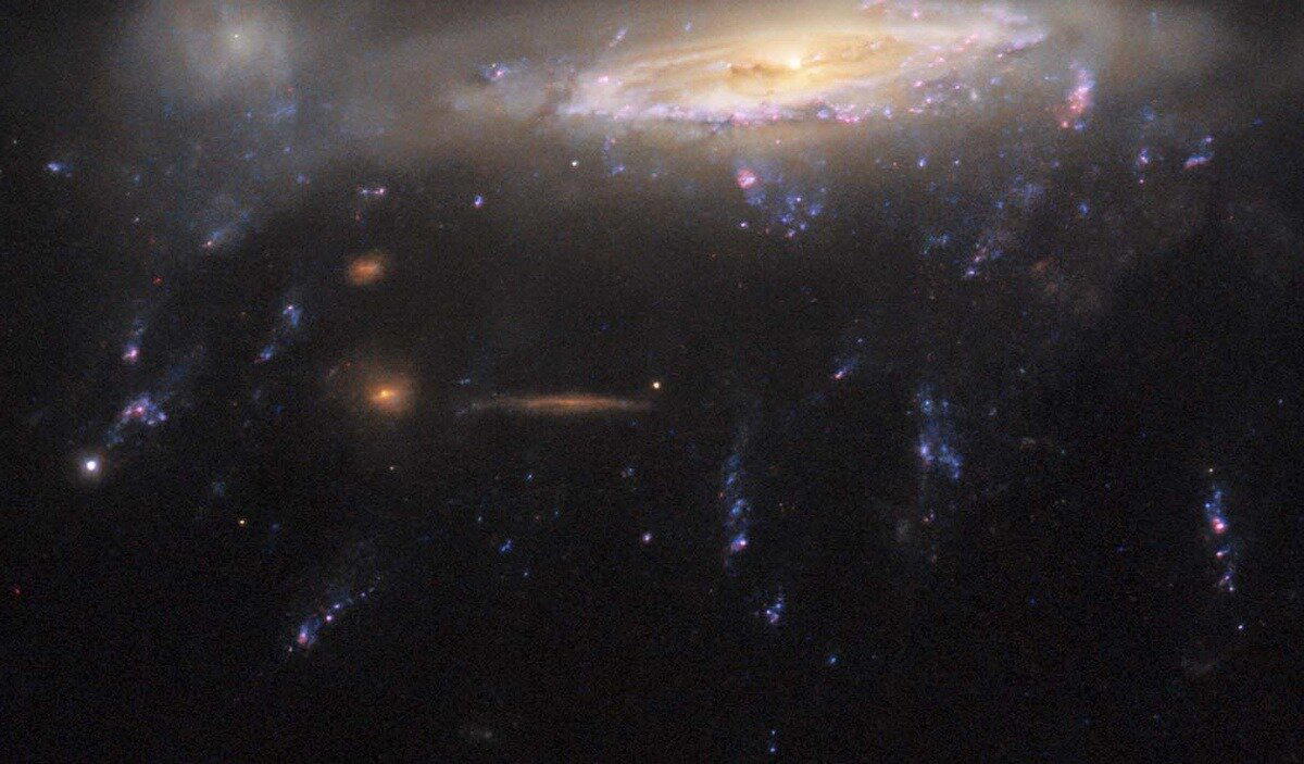 За "щупальцами" видны фоновые галактики, которые выглядят красно-коричневыми из-за высокой концентрации пыли, выдуваемой из PGC 29820 / © NASA/ESA